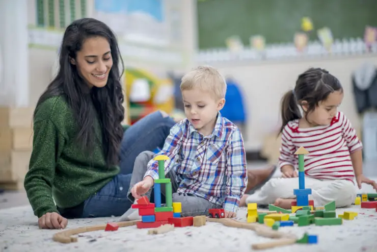 Un niño y una niña de preescolar están adentro con su maestra. Están sentados en el suelo y jugando juntos con coloridos bloques.
